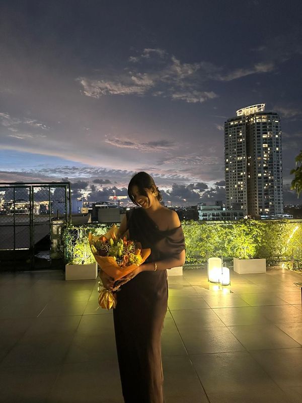 Samadei on a rooftop at blue hour, holding a bouquet, the Colombo skyline behind her.